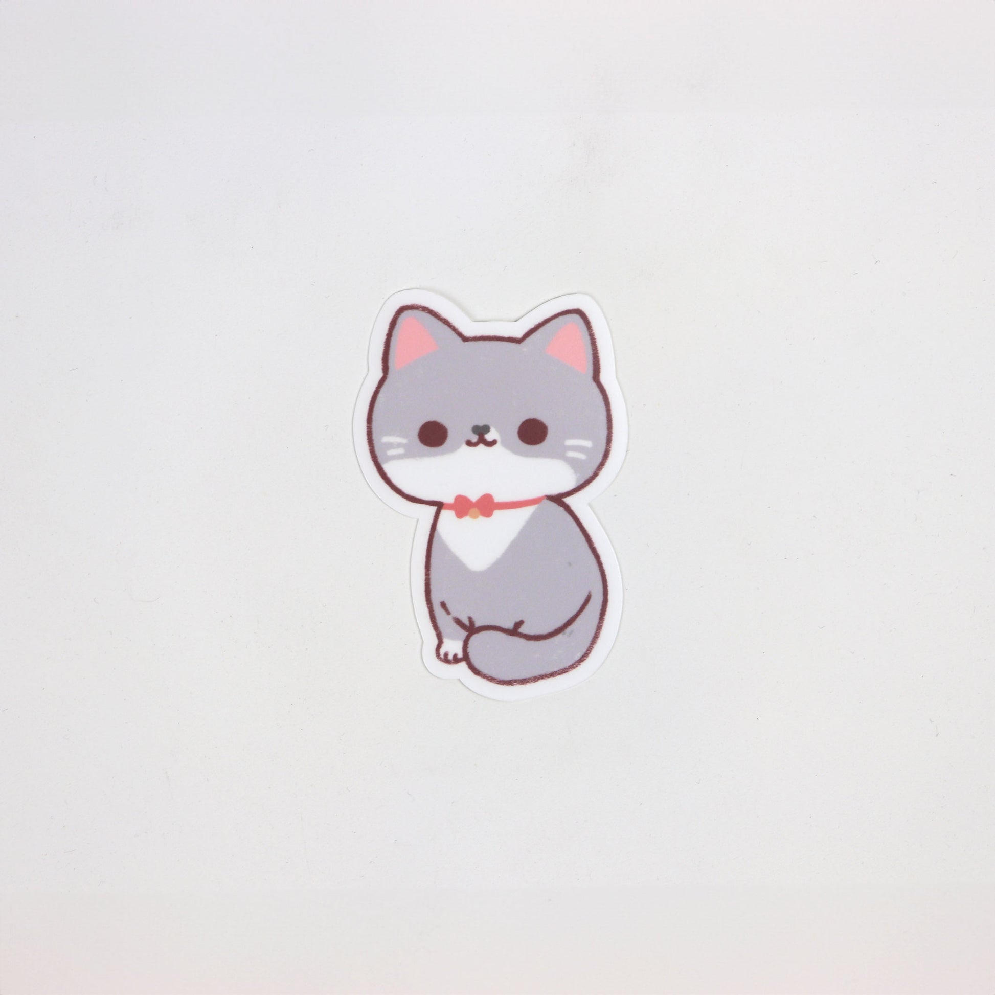Matte die-cut waterproof sticker of a sitting grey and white cat wearing a red bowtie, approx. 5cm x 7cm.