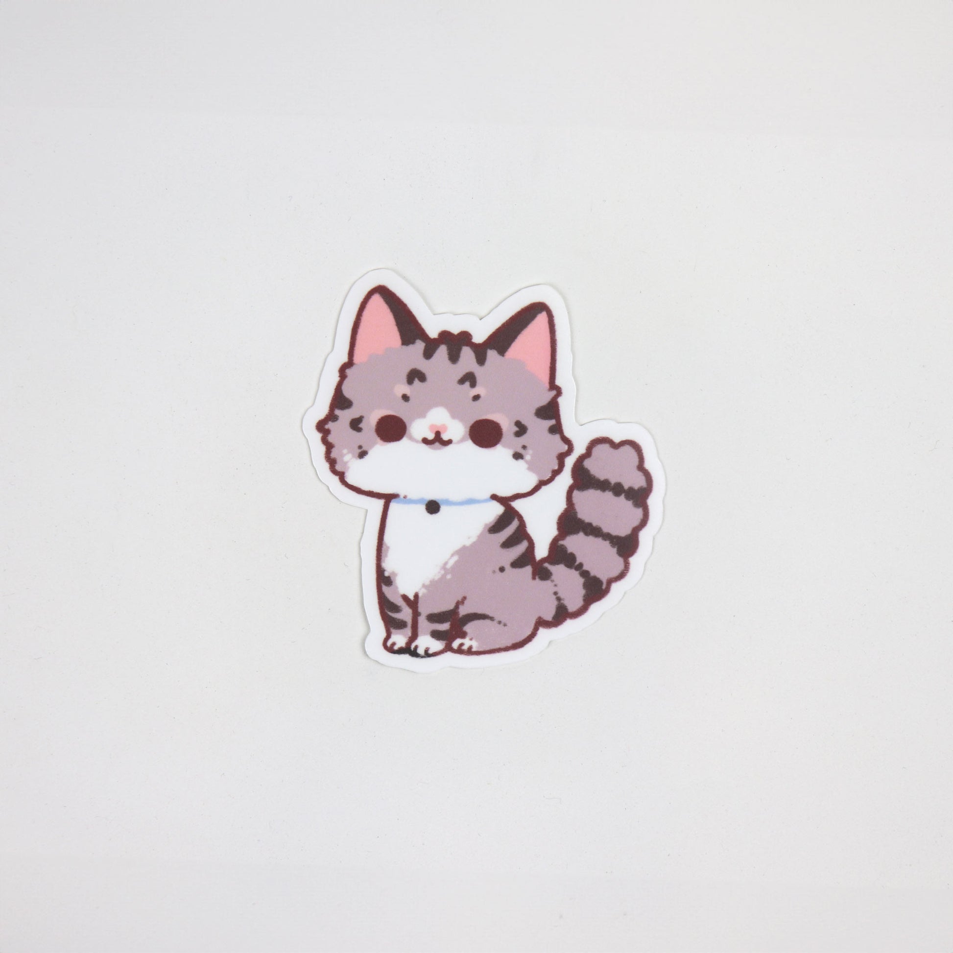 Matte waterproof die-cut sticker of a fluffy tabby cat with grey stripes, blue collar, and pink ears, approx. 5cm x 7cm.